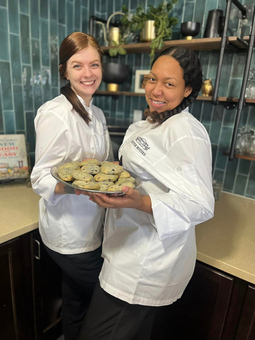 Two smiling female chefs in white uniforms proudly display a plate of freshly baked cookies in a modern kitchen with green tile backsplash.