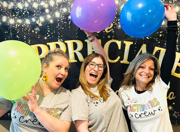 Three enthusiastic women in 'Activities Crew' shirts holding colorful balloons, celebrating in front of an 'Appreciate' banner.