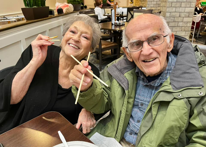 A cheerful elderly couple at a dining table, happily posing with chopsticks. The woman in black and the man in a green jacket share a joyful moment, reflecting the warmth of the community.