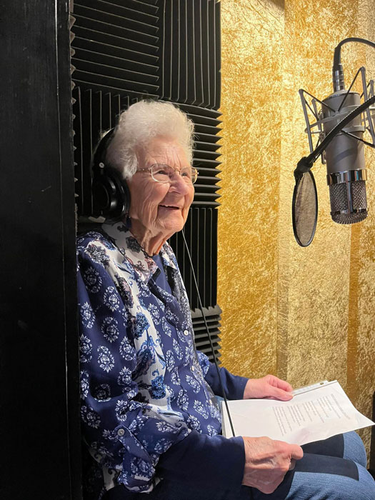 An elderly woman in a soundproof recording booth, wearing headphones and holding a script. She smiles warmly, dressed in a blue floral blouse, ready to record a holiday song.