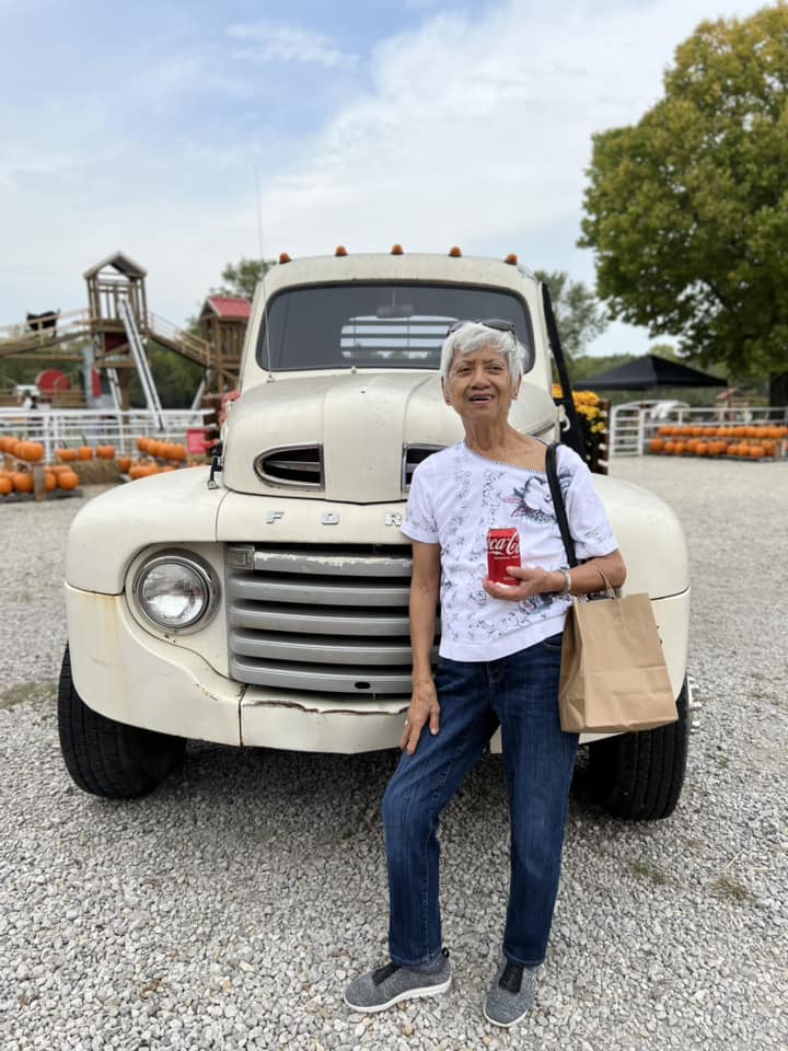 An elderly woman stands in front of a vintage white Ford truck, holding a can of Coca-Cola and a paper bag. She is dressed casually in a white patterned t-shirt, blue jeans, and gray slip-on shoes, enjoying a sunny day at what appears to be a pumpkin patch.