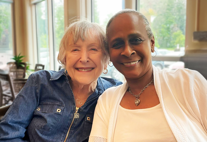 Two elderly women sitting close together, smiling warmly and enjoying each other's company in a bright and cozy room.