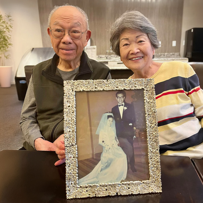 Elderly couple smiling and holding a framed wedding photo, celebrating their lasting love and many years of marriage.