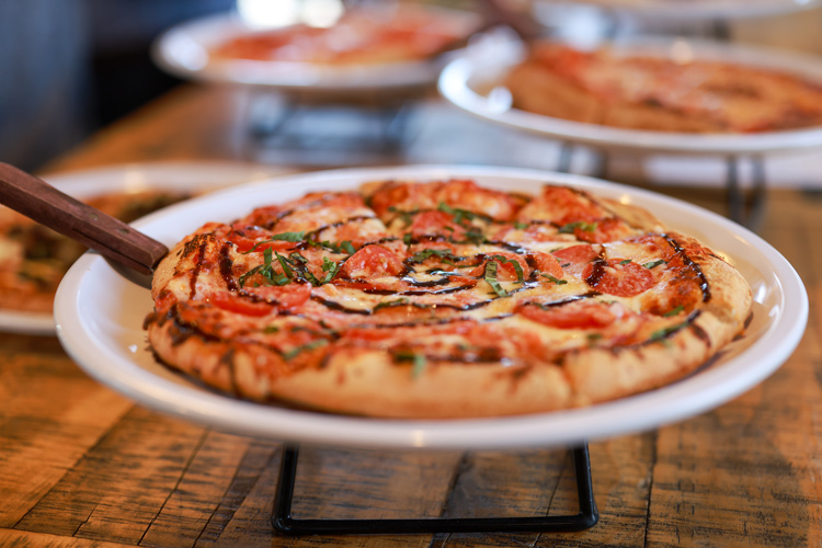 A freshly baked pizza topped with pepperoni, cheese, and a drizzle of balsamic glaze is presented on a white plate, sitting on a wooden table. In the background, more pizzas can be seen, highlighting a delightful spread ready to be enjoyed.