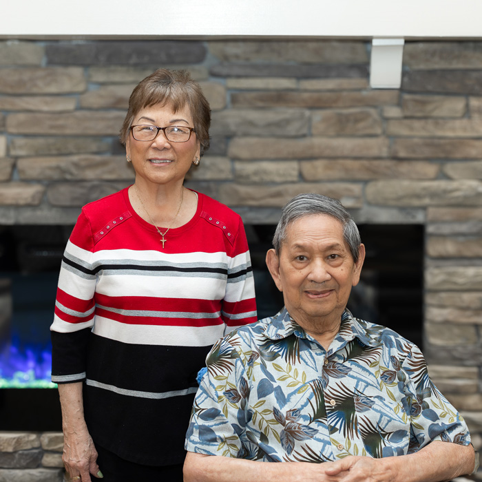 n elderly couple poses in front of a stone fireplace. The woman, standing, wears glasses and a red, white, and black striped sweater. The man, sitting, has a slight smile and wears a blue and white floral shirt. They both look content and happy together.