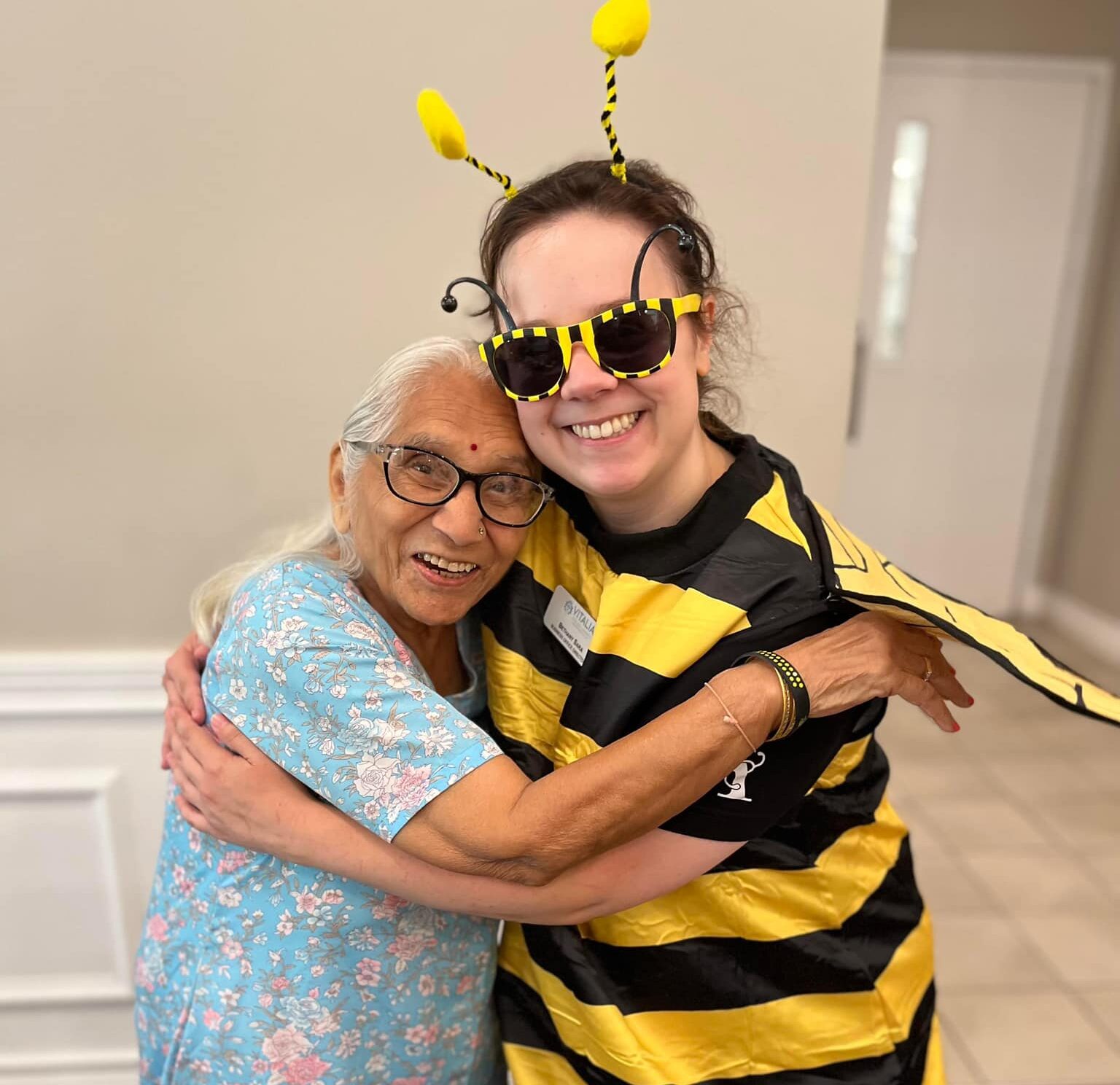 Elderly woman in a blue floral dress joyfully hugging a young woman dressed in a bee costume, both smiling brightly.