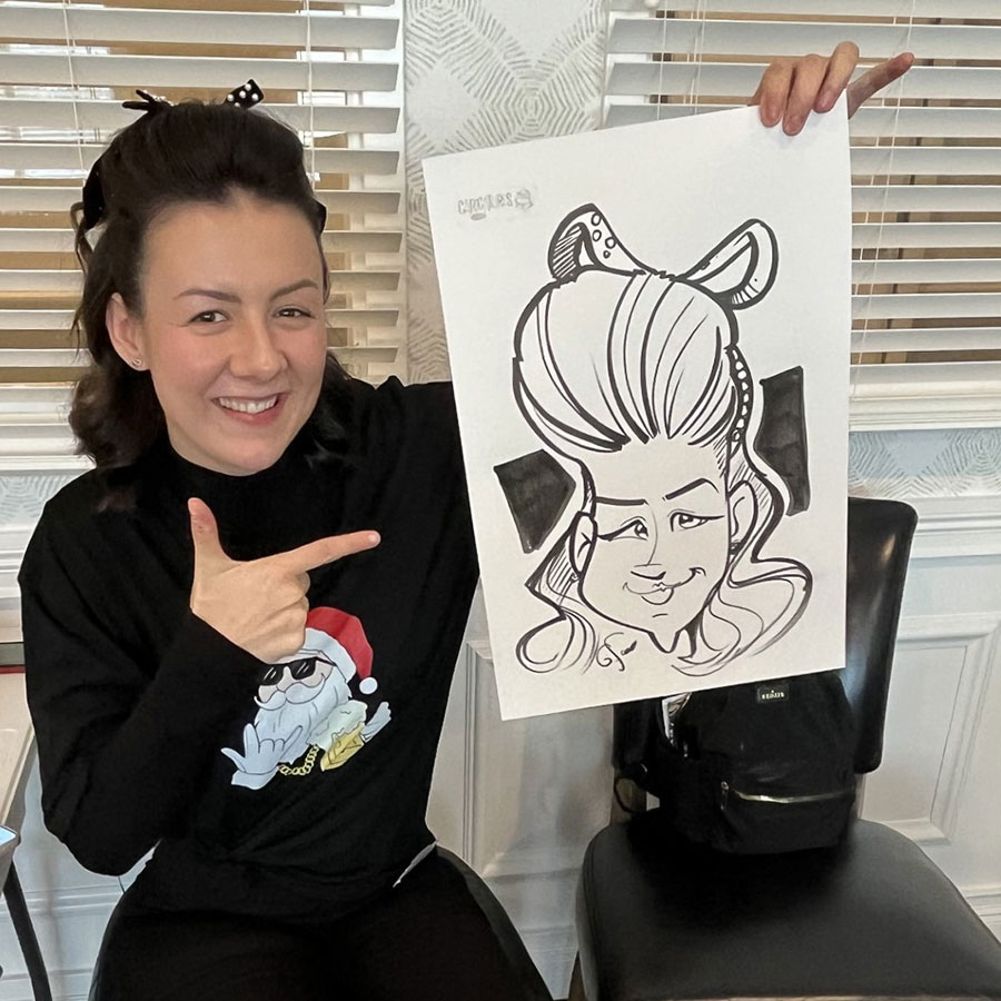 A woman is sitting indoors, smiling and pointing at a caricature drawing of herself. She is wearing a black turtleneck sweater with a Christmas-themed design featuring a Santa Claus. Her hair is styled with a bow, matching the caricature. The atmosphere is cheerful and lively, with the woman proudly showcasing the artistic representation.