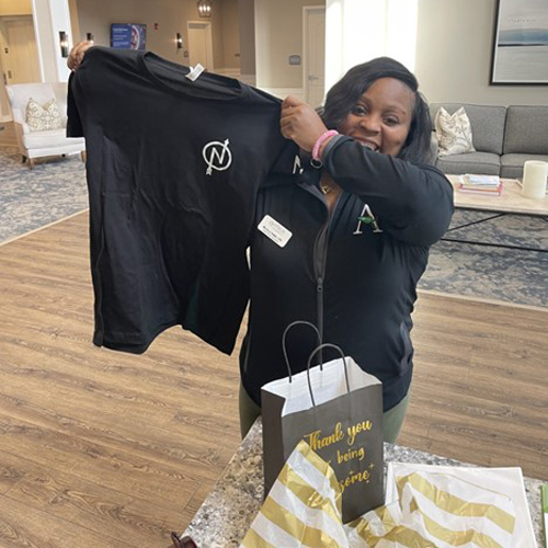 Smiling woman in a black jacket proudly holding up a black T-shirt with a logo, with gift bags and a 'Thank you' card on the table.