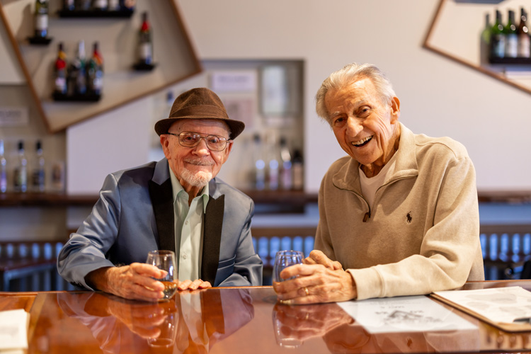 Two elderly men sitting at a bar, smiling and enjoying drinks together, creating a warm and friendly atmosphere.