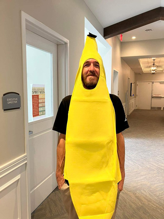 Man in a bright yellow banana costume smiling in a hallway with white walls and doors, participating in a fun event.