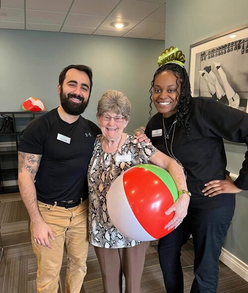 Smiling senior resident holding a beach ball stands between two staff members, one male and one female, during a festive indoor event, with all three posing happily together.