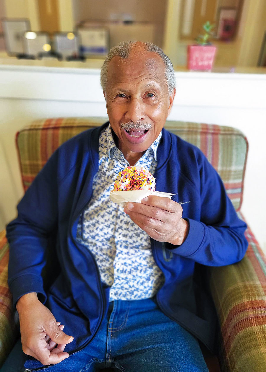 Smiling senior resident in a blue jacket and floral shirt enjoys a sprinkled donut while sitting comfortably on a plaid chair, capturing a moment of joy and indulgence.