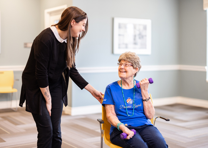 Smiling senior senior resident in a blue shirt exercises with a purple dumbbell while seated, receiving encouragement from an employee standing beside her in a fitness room.