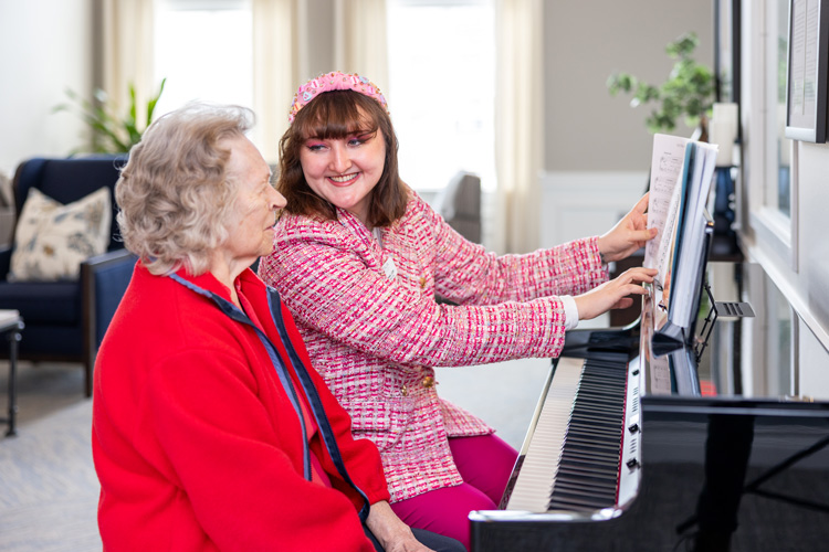 A woman in a pink jacket and matching headband smiles while showing piano sheet music to an senior resident in a red sweater, both sitting at a grand piano in a bright room.