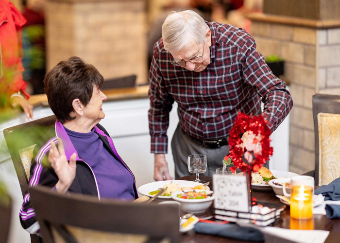 Senior residents enjoy a lively conversation while sharing a meal in a decorated dining area. The table is set with colorful salads, dishes, and a festive red centerpiece.