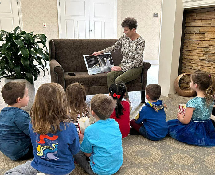 A senior resident reads a picture book to a group of attentive young children seated on the floor in front of a couch, during a storytime session in a cozy, well-lit room.