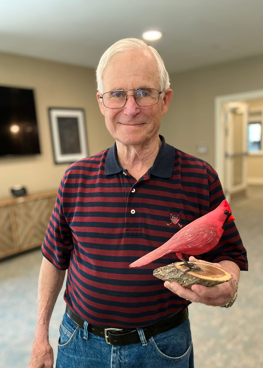 A senior resident with white hair and glasses, wearing a striped polo shirt, proudly holds a detailed wooden carving of a red cardinal, smiling warmly at the camera.