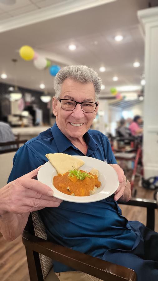 Senior resident smiling while holding a bowl of food topped with a rich orange sauce and garnished with fresh herbs, seated in a brightly decorated dining area.