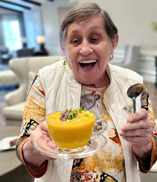Smiling senior resident joyfully holds a bowl of vibrant orange soup garnished with greens, wearing a colorful butterfly-patterned shirt and white vest, ready to enjoy the meal.