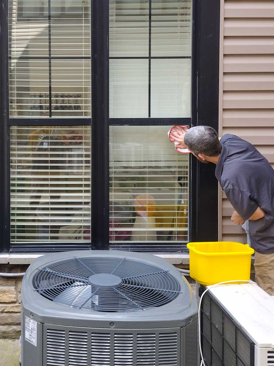 Maintenance worker in a gray shirt cleans the outside of a window near an air conditioning unit, ensuring the senior living community's environment remains clean and well-maintained.