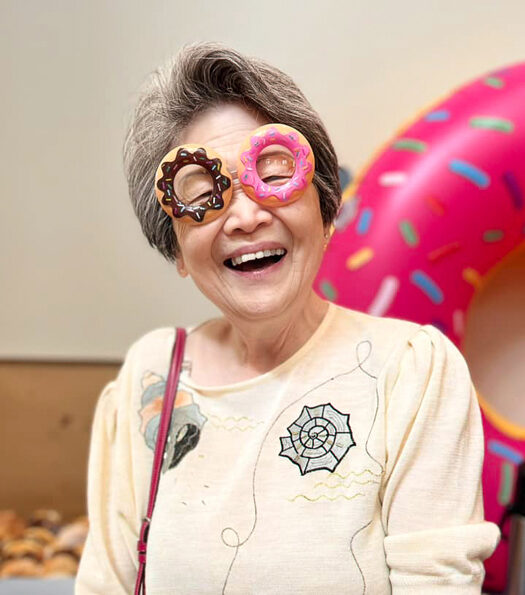 Senior resident joyfully wears playful donut-shaped glasses, standing in front of a large inflatable donut with sprinkles, highlighting a fun and lighthearted atmosphere.