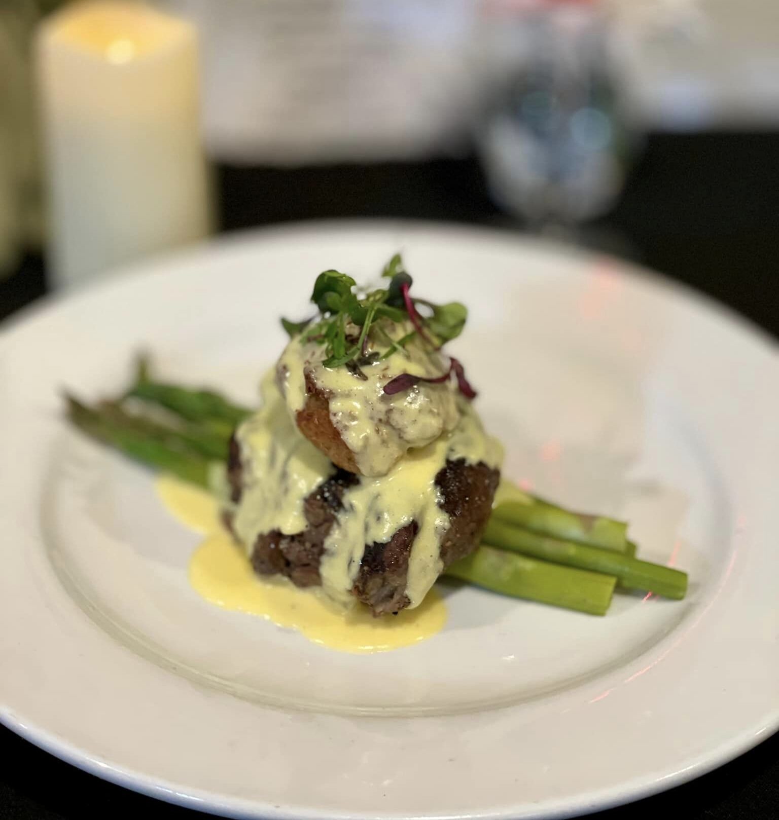 Plated gourmet steak with asparagus, topped with creamy hollandaise sauce and garnished with microgreens, set against a blurred background of a candle, flowers, and drink.
