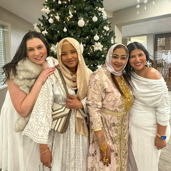 Four women in elegant attire standing together in front of a decorated Christmas tree, celebrating diversity and holiday traditions in a senior living community.