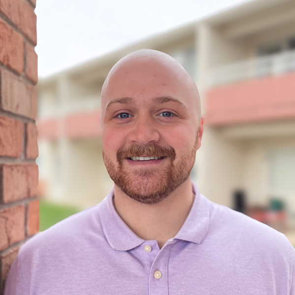 Carter Daniel, Senior Living Director at Walden Place Senior Living, smiling in a light purple polo shirt, stands outside near a brick pillar and residential building.