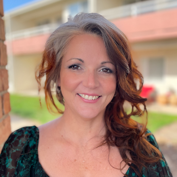 Holly Sindelar, Executive Director at Walden Place Senior Living, smiles warmly in a green patterned top with a soft outdoor backdrop of the community behind her.