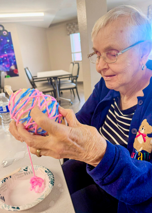 Smiling senior resident wraps colorful yarn around a balloon during a springtime craft activity at a senior living community.
