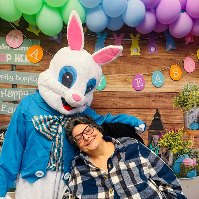 Senior woman happily poses with the Easter Bunny mascot in front of a festive spring-themed Easter display with balloons and colorful signs.