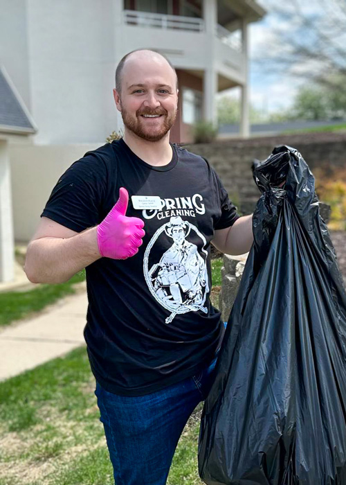 Team member wearing a “Spring Cleaning” shirt gives a thumbs up while holding a full trash bag outdoors at a senior living community.