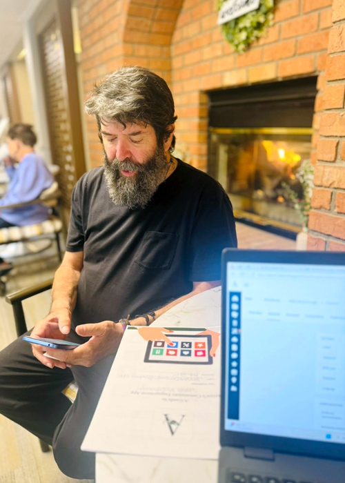 A senior man in a black t-shirt sits by a fireplace using a smartphone, with a laptop open nearby showing the Arrow app training materials.