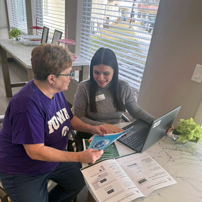 A senior woman in an Iowa t-shirt reviews printed materials about the Arrow app with a smiling team member using a laptop in a bright, sunlit room.