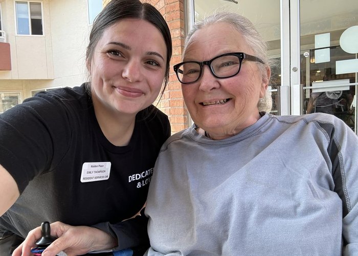 Walden Place Senior Living employee in a black T-shirt take a selfie with a senior resident outside of of the building on a sunny day.