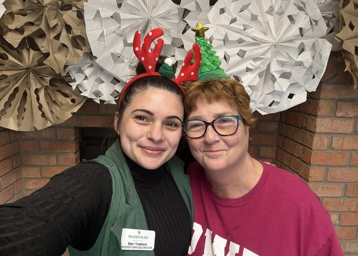 Walden Place Senior Living's Resident Services Director wearing a black sweater and green vest and antler head band posing close to a resident with Christmas tree headband pose together in front of paper snowflake decor.