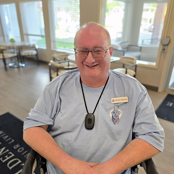 Smiling man wearing glasses and a light blue shirt sits in the lobby of a senior living community.