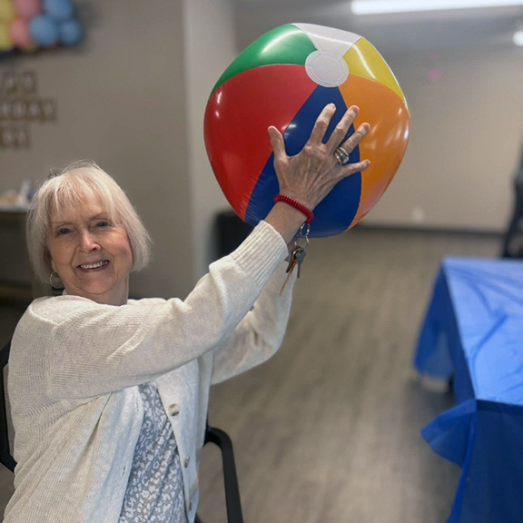 Woman happily lifts a colorful beach ball during a senior living game, enjoying lighthearted fun, laughter, and wellness.