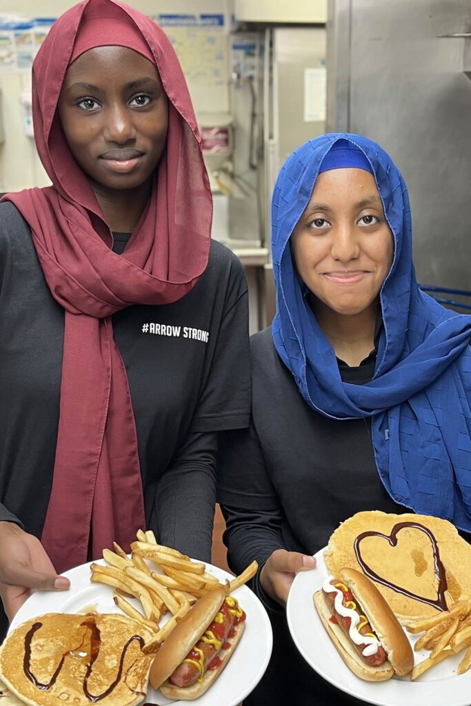 Two team members in #ArrowStrong shirts smile while holding plates with hot dogs, fries, and pancakes decorated with syrup designs in the kitchen.