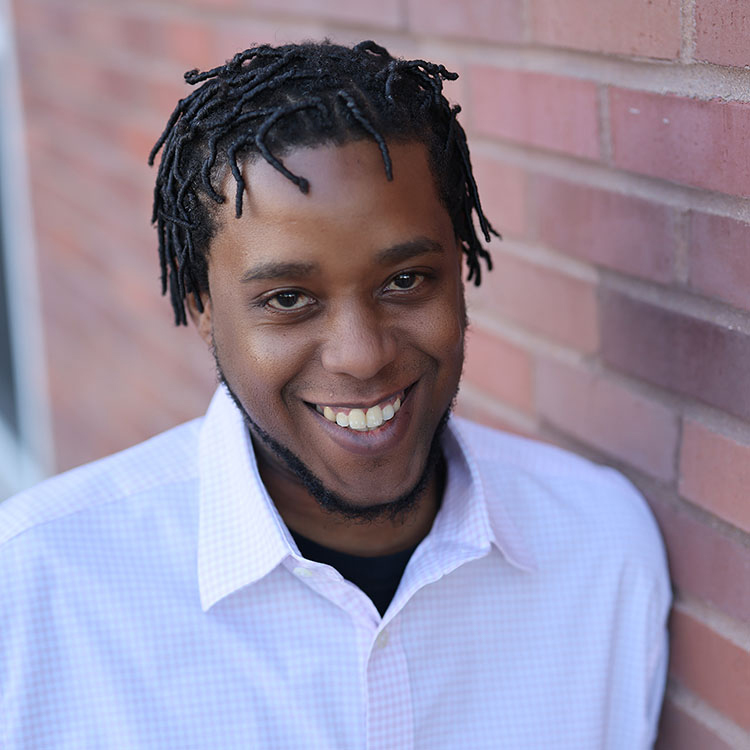 Ken Duff, Interim Executive Director at Walden Place Senior Living, smiling in a headshot. He has short twists and a trimmed beard, wearing a light checkered shirt while leaning against a brick wall.