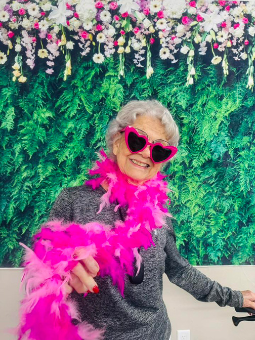 Smiling senior woman posts in front of a flowered backdrop with a hot pink feather boa, pink heart sunglasses and freshly painted fingernails.