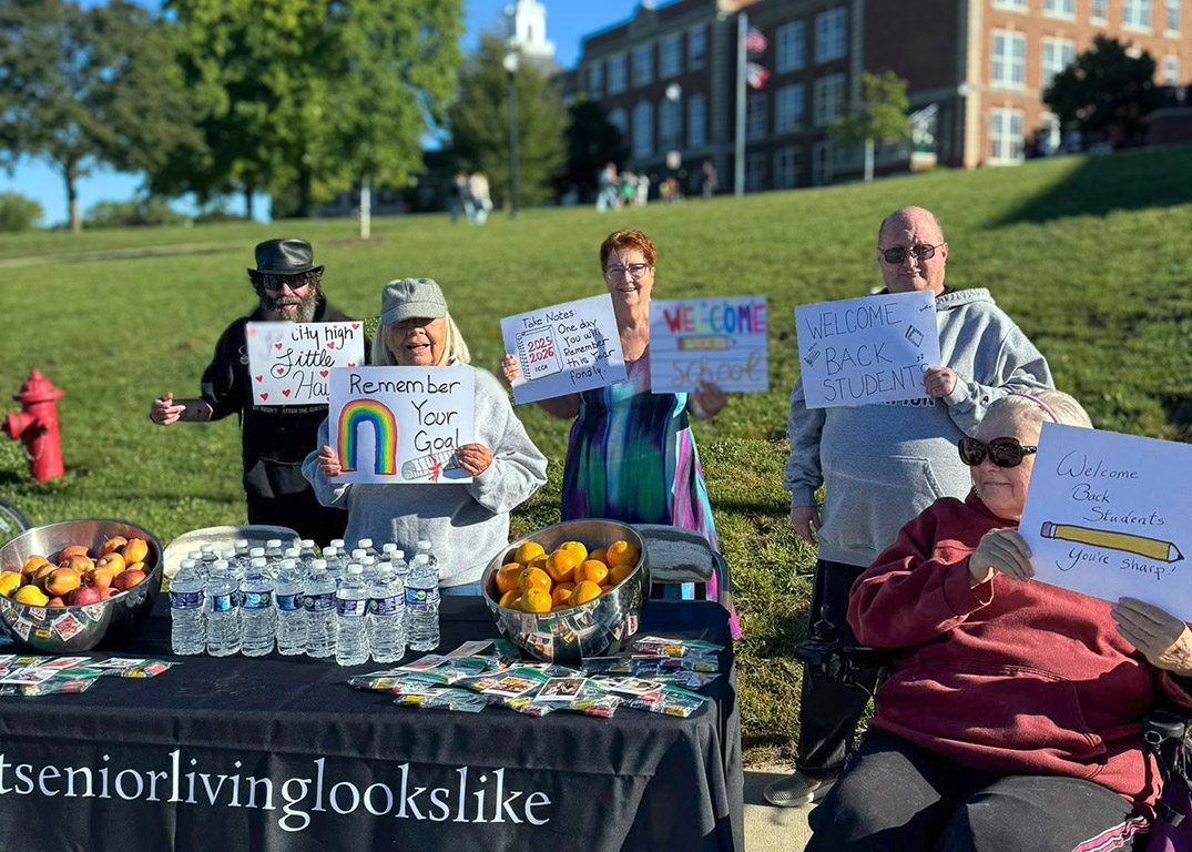 A group of seniors hold up encouraging signs for returning students, manning a table full of healthy snacks, treat bags and water.