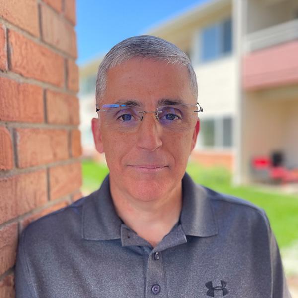 Jeff Steadman, Plant Operations Director at Walden Place Senior Living, standing outdoors near a brick wall with a light-colored building in the background. He wears glasses and a dark gray collared Under Armour shirt, smiling slightly in natural sunlight.