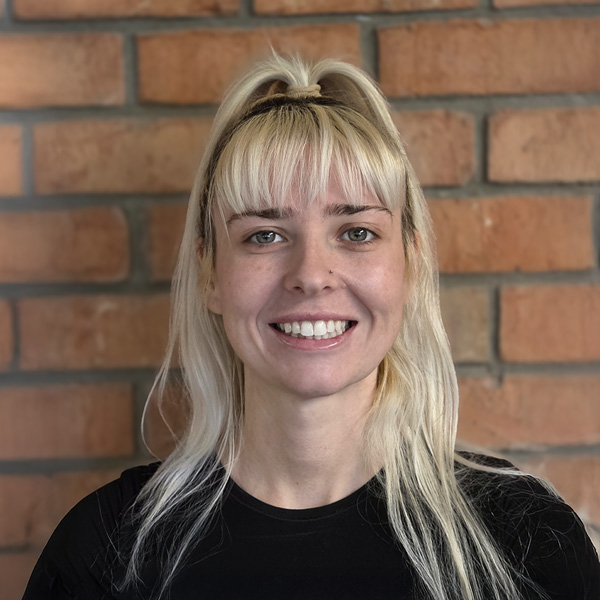 Kristen Hawkins-Hamer, Business Operations Director at Walden Place Senior Living, smiling in front of a warm brick wall while wearing a black top with her blonde hair styled in a half ponytail.