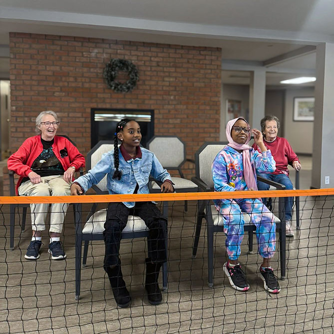 Senior residents and visiting youth smile beside a beachball volleyball net.
