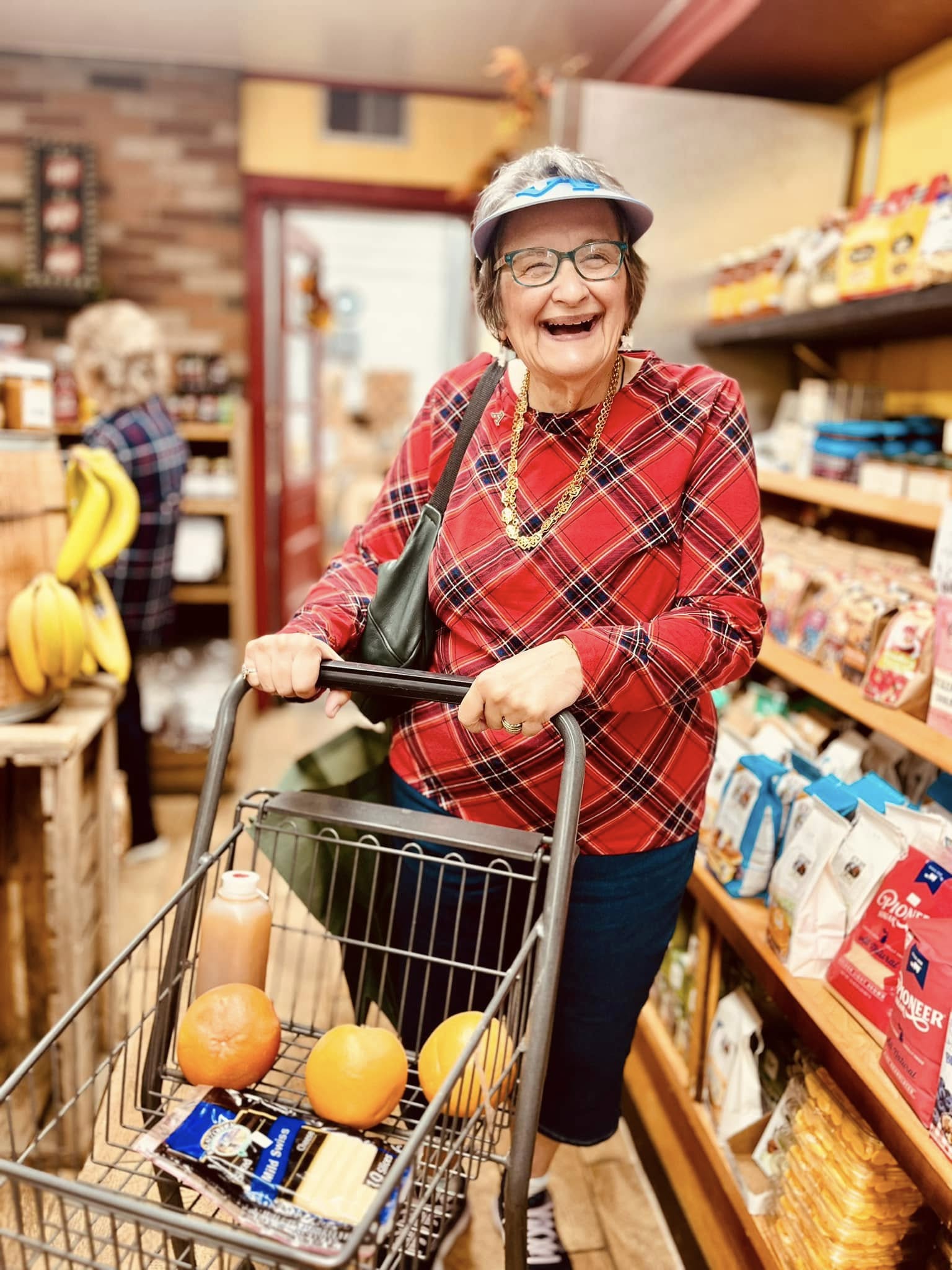 A senior living resident, a woman in a visor, necklace, and plaid shirt, laughs joyfully while pushing a grocery cart during shopping.