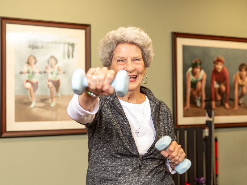 Senior resident smiling while lifting light hand weights during a fitness class, staying active and engaged in a welcoming senior living community exercise space.
