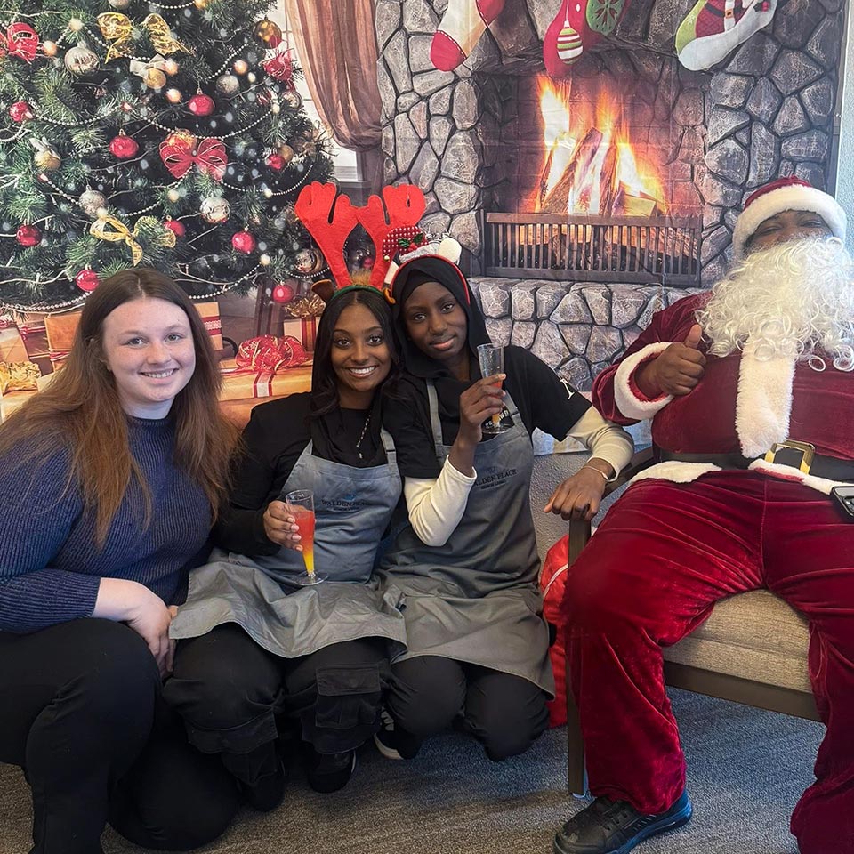 Three Walden Place team members smile alongside Santa Claus in front of a richly colored Christmas backdrop.