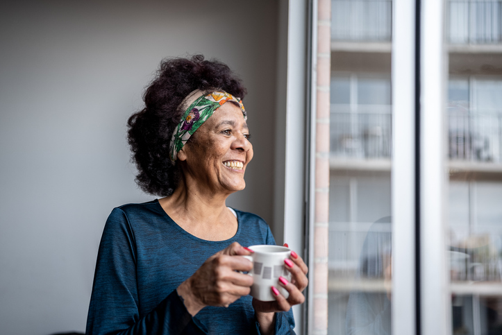 Senior woman with her hair pulled back by a colorful headband enjoys a morning coffee while looking out the window of her senior living apartment.
