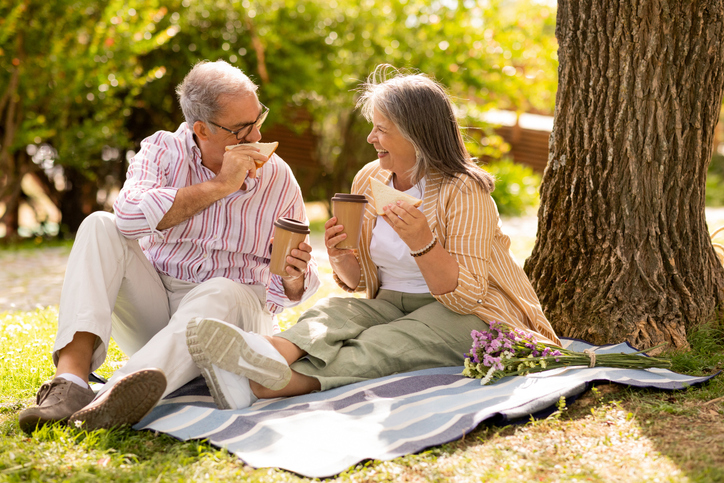 A senior couple sits on a blanket under a tree while enjoying a coffee together, one of them, a woman, has a bouquet laying next to her.